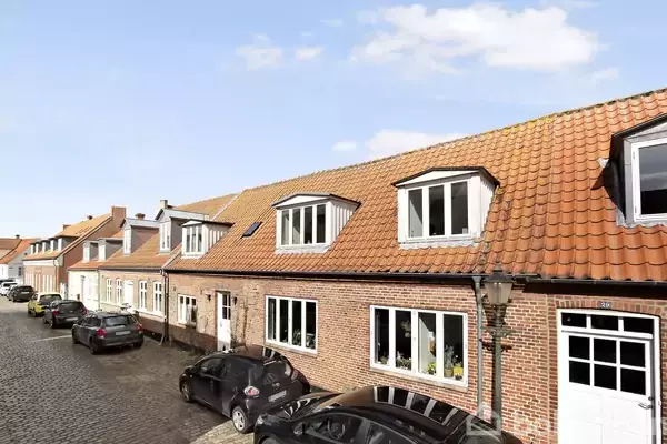 Rækkehuse med røde tegltage står langs en brostensbelagt gade. Flere sorte biler er parkeret foran husene, og himlen er blå med få skyer.