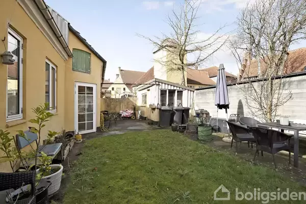 Gårdhave med græsplæne omkranset af et gult hus og et hegn. Der står borde, stole og en parasol i haven, omgivet af forårsplanter under en blå, klar himmel.