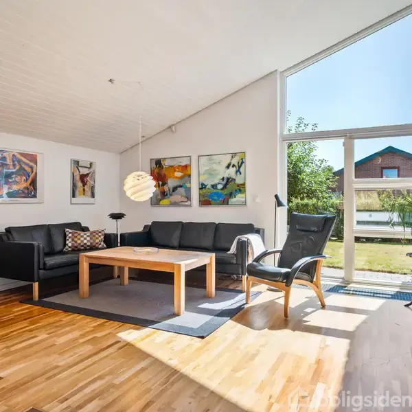 En stue med sorte møbler og et sofabord står i centrum. På væggene hænger farverige malerier. Store vinduer til højre viser en have og naboens hus.