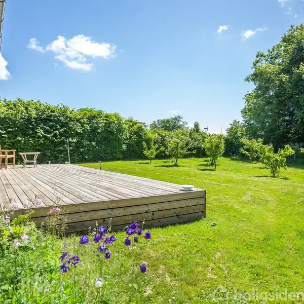 En træterrasse med stole og bord står ved siden af et hus. Terrassen er omgivet af en frodig græsplæne og grønne træer under en klar, blå himmel.
