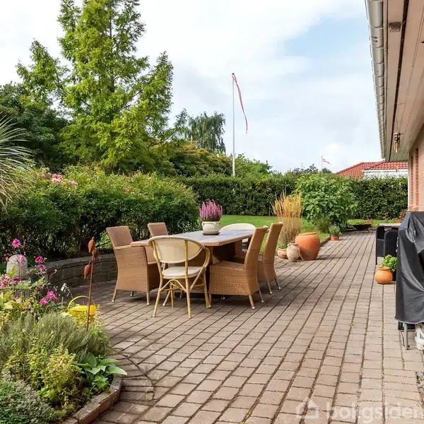 En udendørs terrasse med spisested inkluderer bord og seks stole. Haven omkring er frodig, med blomster, buske og træer. En rød murstensbygning ligger til højre for terrassen.