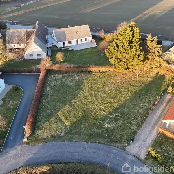 Et åbent grønt græsareal er omgivet af huse med røde tage langs en snoet vej i et landdistrikt. Baggrunden viser flere marker og træer.