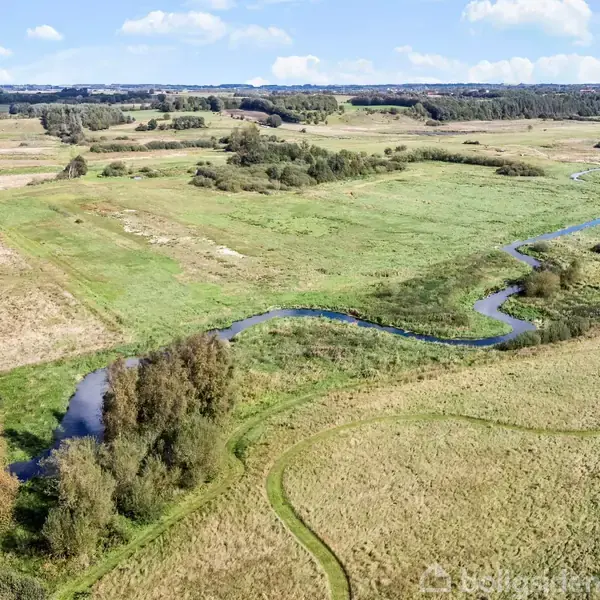 Flod slynger sig gennem et åbent landskab med grønne enge og spredte træer under en blå himmel med skyer.