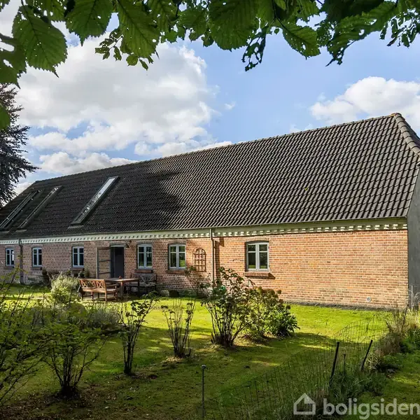 Hus bygget i sten med sort tag, beliggende i en grøn have med buske og træer. Sollys skinner gennem træernes blade, og himlen er let overskyet.