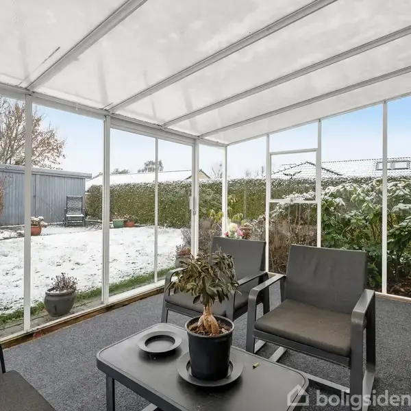 Overdækket terrasse med sorte havemøbler står foran et vindue ud mod en sneklædt have. En potteplante er placeret på bordet. Haven er omkranset af grønne buske og et skur.