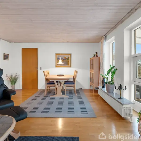 En spisestue med et bord og fire stole står på et tæppe. Til venstre er en sort lænestol. Vinduer til højre giver naturligt lys. Indretningen er minimalistisk.