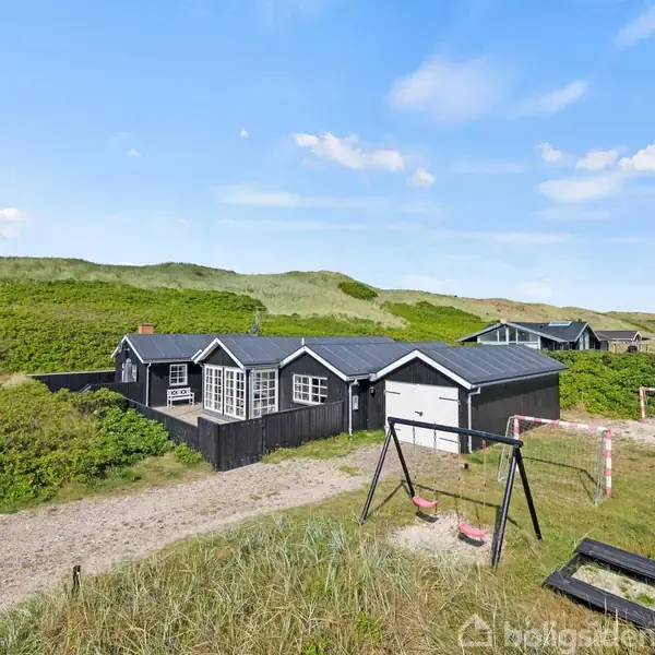 Et sort sommerhus står blandt grønne klitter under en blå himmel. En gyngestativ og fodboldmål er placeret i sandet foran hovedbygningen.