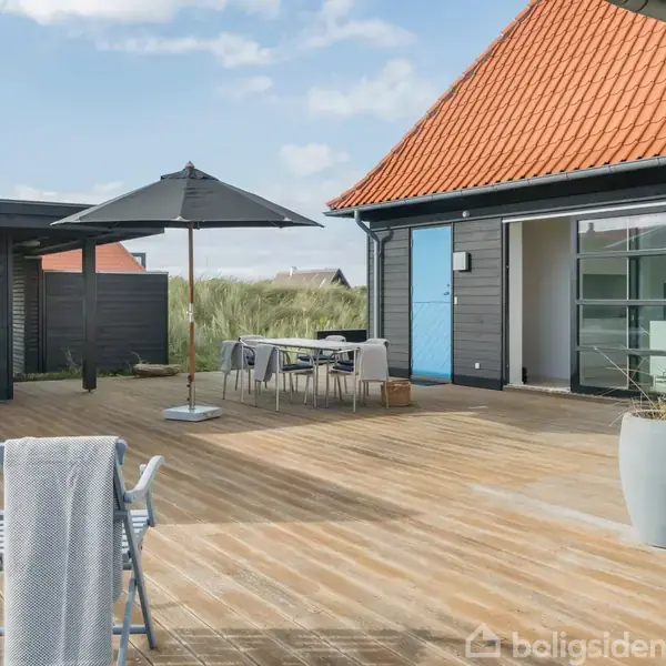 Terrasse med havemøbler og parasol, der står på et trædæk foran et sort-hvidt hus med rød tag. En gasgrill står i hjørnet, og udsigten inkluderer græs og en klar himmel.