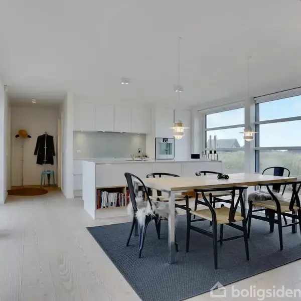 Et spisebord med stole står på et tæppe, omgivet af store vinduer i en lys og moderne spisestue. Køkkenet er i baggrunden, med trappe til højre.