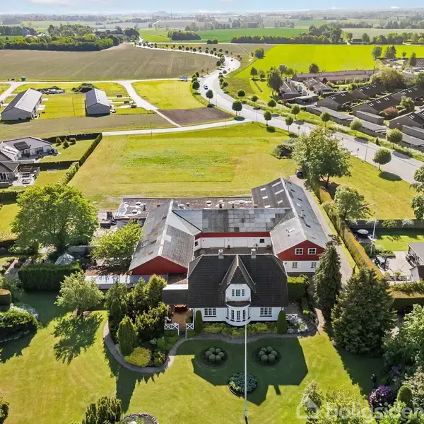 Stor villa med mørkt tag set oppefra, omgivet af grønne plæner og træer. Husene ligger spredt i et landligt landskab med marker og veje i baggrunden.