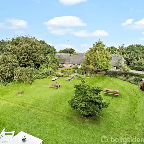 Grøn græsplæne med picnicborde og en enkelt busk. I baggrunden ses et traditionelt hus omgivet af træer. Serien af skyfrie blå himmel tilføjer til den idylliske atmosfære.