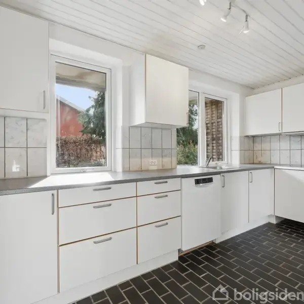 Hvidt køkken med flisegulv og sorte fuger. Skabe og skuffer er i minimalistisk design. To vinduer giver naturligt lys. Mosaik-vægbeklædning skaber kontrast.