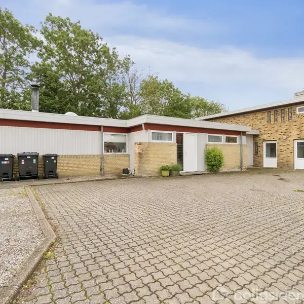 Bygning i én etage med fladt tag står på flisebelagt grund. Flere skraldespande er placeret langs væggen. I baggrunden ses træer mod en klar himmel.