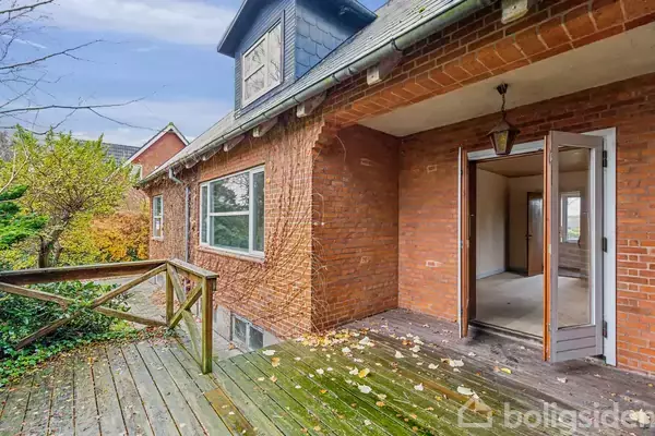 En murstensbygning med åben dør til et rum indenfor. En træterrasse med lidt visne blade. Omgivet af nogle træer og buske.