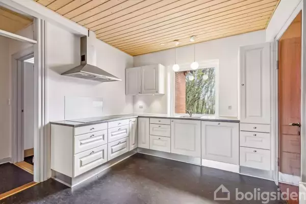 Hvidt køkken med grå skabslåger, stålvask og emhætte. L-formet arbejdsområde. Vindue med udsigt til træer. Loft med træpaneler og tre kugleformede lamper hængende over vinduet.