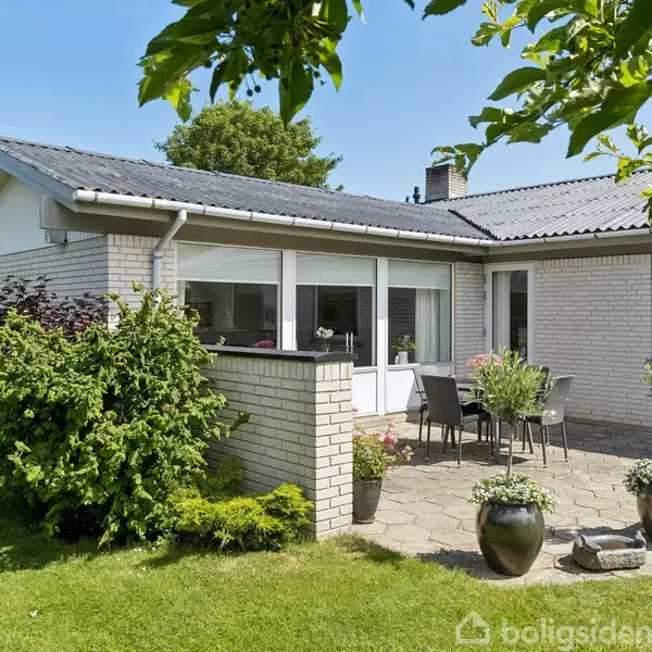 En hvid murstensbungalow har en solrig terrasse med havemøbler. Grønne buske og græsplæne omkranser huset. Blå himmel og træer i baggrunden skaber en rolig haveatmosfære.