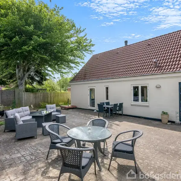 Et hus med rødt tegltag og hvid facade har en terrasse med havemøbler. I baggrunden er der hegn og et stort træ. Til venstre står et sofaarrangement.