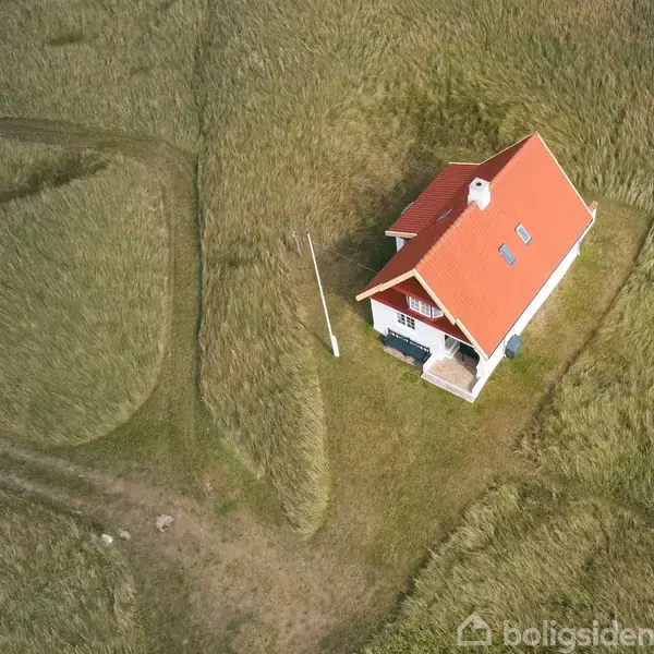 Rødt taghus står midt i græsklædt klitlandskab. Huset er omgivet af stier og sandede områder, skaber en følelse af isolation og ro.