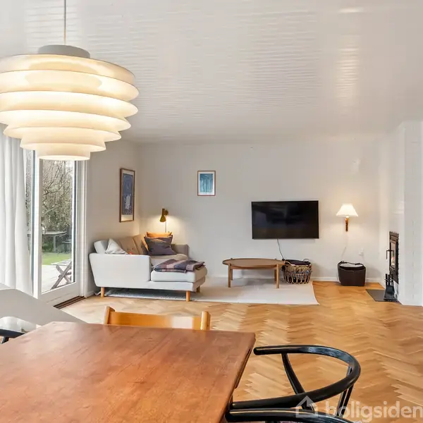 En stue med en sofa og et væghængt TV. Rummet har trægulv og åbner ud mod en terrasse gennem store vinduer. Lamper belyser hjørnet, og en bogreol står til højre.