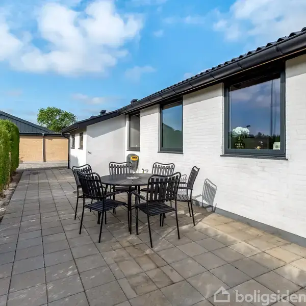 Et havesæt med sorte stole og bord står på en flisebelagt terrasse ved en hvid murstensbygning. Ved siden af hækken er en asfalteret sti, og en garage ses i baggrunden.