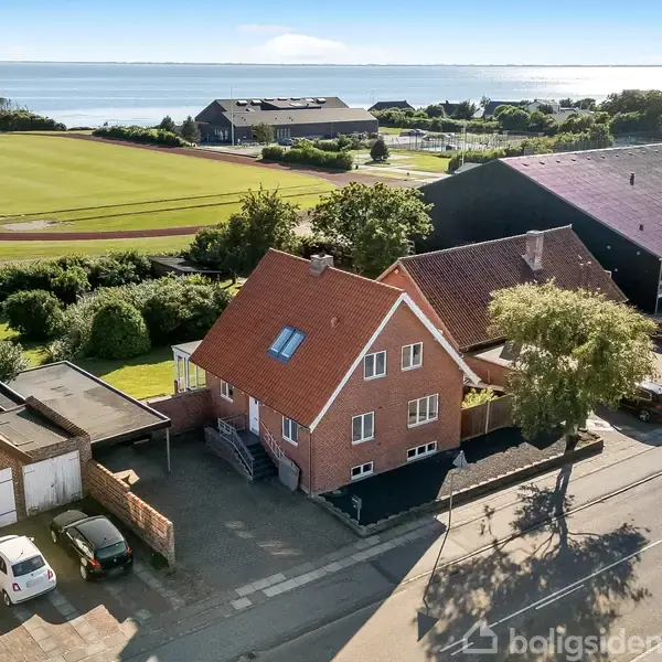 Et rødt murstenshus med skråtag står ved siden af en vej. Bagved ses en sportsbane og derudover havet i horisonten. To biler holder parkeret nær huset.