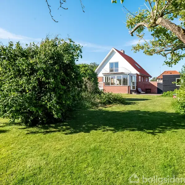 Et hus med en rød tag og stor glasveranda står i en grøn have med græsplæne, buske og træer under en klar blå himmel.