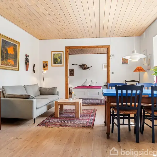 Sofa står ved siden af et lille bord med lampe, i en stue med træloft. Spisebord med blå stole er til højre nær vindue. Soveværelse ses gennem åben dør.