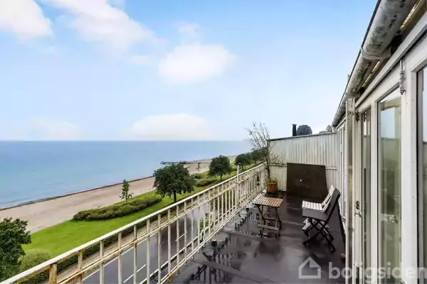 Altan med borde og stole vender ud mod havet, hvor stranden og træer strækker sig langs kystlinjen. Skyfri blå himmel dækker landskabet.