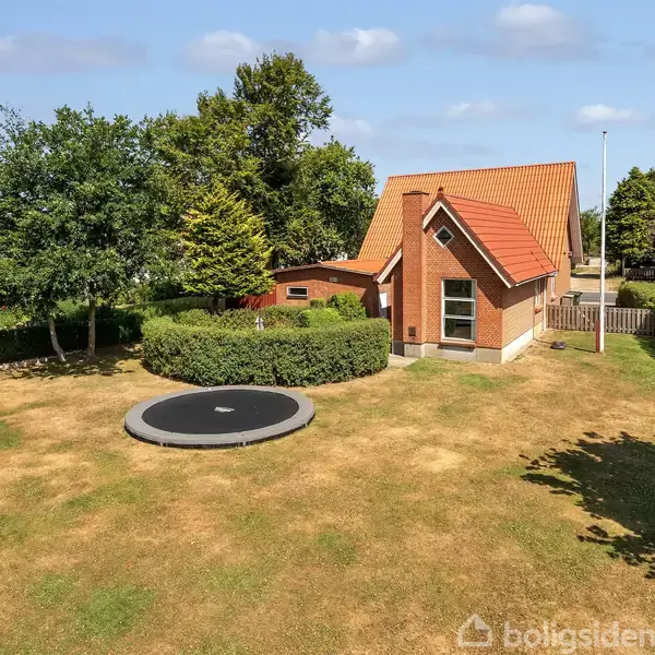 Et rødt murstenshus med skråt tag står i en velplejet have med en trampolin i græsset. Omgivet af hække og træer under en blå himmel.