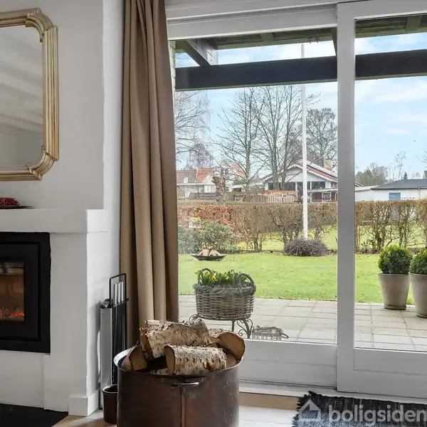 En pejs med levende ild står i stuen med træbrændsel foran. Store vinduer viser en terrasse og en have med buske. En spejl hænger over pejsen.