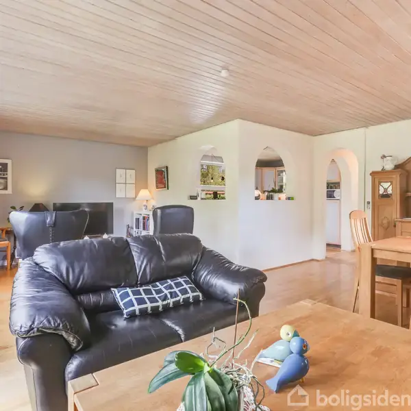 En stue med sorte lædersofaer, placeret ved et sofabord. Pejs og spisebord med stole i træ ses i samme rum. Store vinduer slipper lys ind.