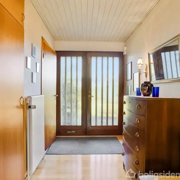 En entré med en kommode til højre og store glasdøre for enden fører ud til det fri. Venstre side har indbyggede skabe. Et spejl hænger over kommoden.