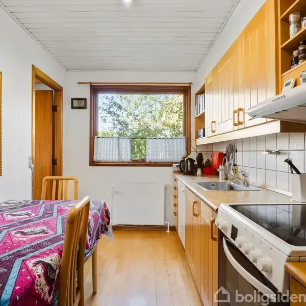 Køkkenbordet er dækket med en farverig dug, mens krydderier står organiseret på hylder over en komfur og vask. Et stort vindue giver udsigt til træer udenfor.