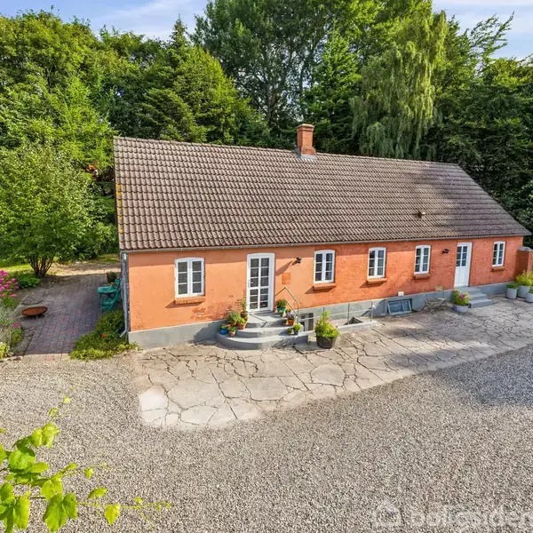 Et orange murstenshus med mørkt tag står på en grusplads. Foran huset er der trapper med blomsterkrukker. Omgivet af grønne træer og buske i en have.