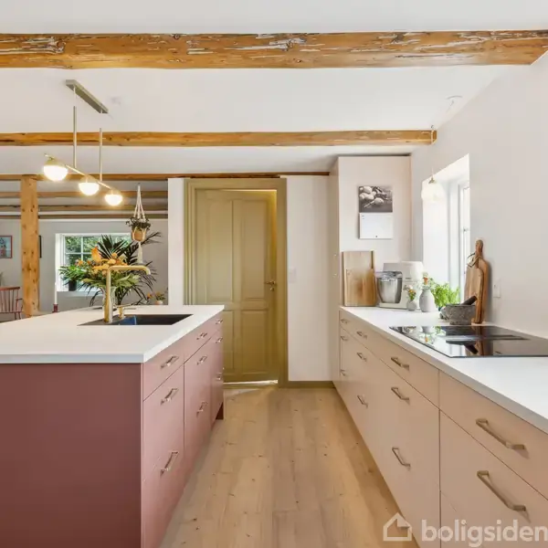 Køkkenbord omgivet af skabe under en loftlampe; rummet har synlige bjælker og en trædør i baggrunden. Dekorationer omfatter planter og billeder.