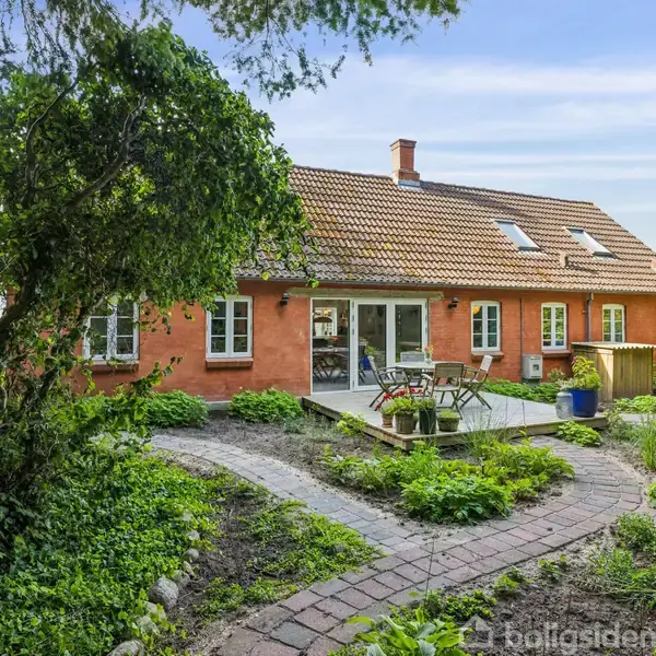 Rødt murstenshus med tagvinduer, omgivet af en frodig have med stenbelagte stier. En træterrasse foran huset har bord og stole, omringet af grønne planter og træer.