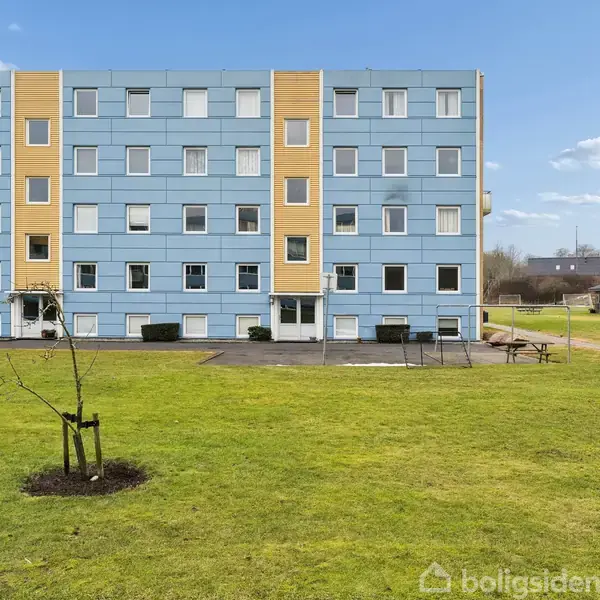 En lys blå og gul lejlighedsbygning i fire etager står i baggrunden. En græsplæne med en nyplantet træ i forgrunden, og en legeplads ses til højre.
