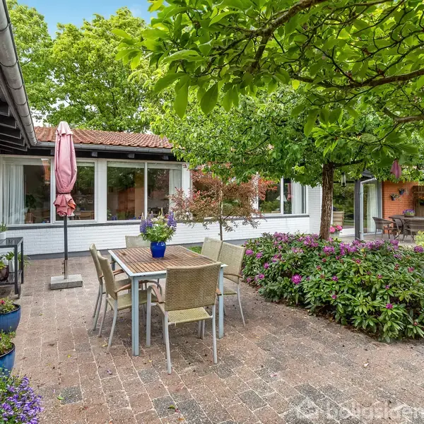 En terrasse med et bord og seks stole under en parasol omgivet af blomsterbede og buske i en frodig have. En murstensvilla ses i baggrunden.