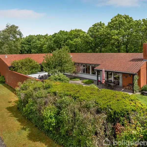 Et stort, rødt murstenshus med skråt tag står blandt frodige grønne træer og buske i en villavej. Haven er velplejet med græs og forskellige planter, grænser op til en stille vej.