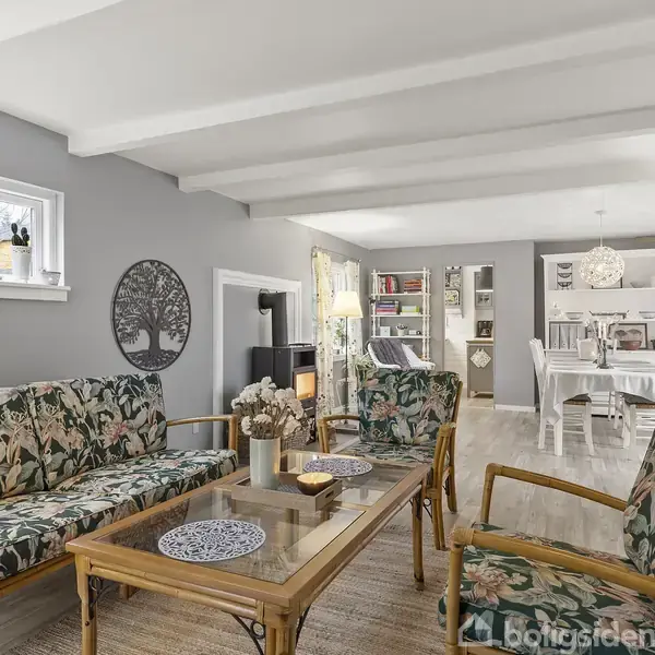 Stue med bambussofa og stole med blomstrede hynder, et sofabord med dekorative tallerkener. I baggrunden ses en spiseplads med hvidt bord og stole, belyst af en lampe.