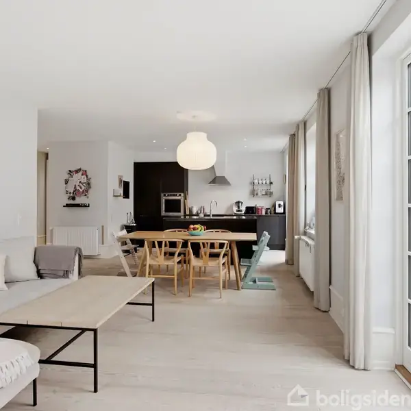Et moderne køkken med grå bordplade og indbygget vask; spisebord med stole ved siden af. I baggrunden ses en stue med sofaer, et vindue og trappen fører opad.