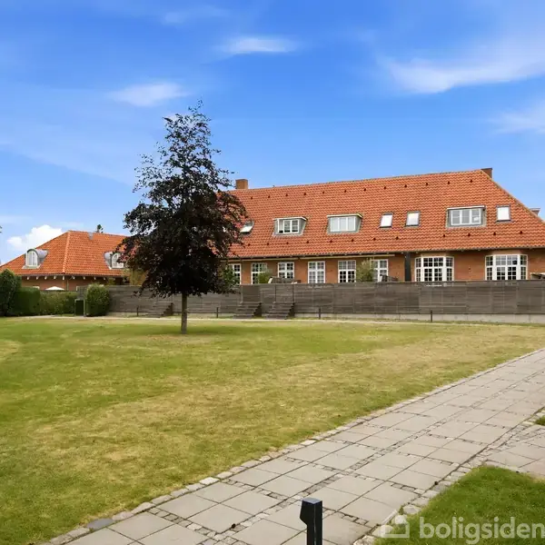 Stue med en sofa og sofabord, et fjernsyn på et TV-bord, to store vinduer med gardiner. Lyse trægulve og vægge. Dekorationer inkluderer vægkunst og planter.