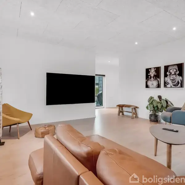 Brun lædersofa står i moderne stue med fladskærm på hvid væg. En gul lænestol står til venstre, og et sofabord er i midten. Billeder og planter dekorerer rummet.