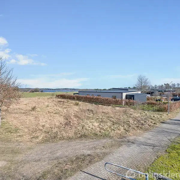 Gruset sti passerer en græsmark ved siden af et boligområde med moderne huse og indkørsler. Der er træer i forgrunden, og i baggrunden ses et åbent landskab og blå himmel.