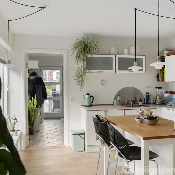Køkkenbord omgivet af barstole står under moderne lamper. Køkkenet har hvide skabe og en plante ovenpå, mens en gang fører ud til en lille entré med hængende jakker.