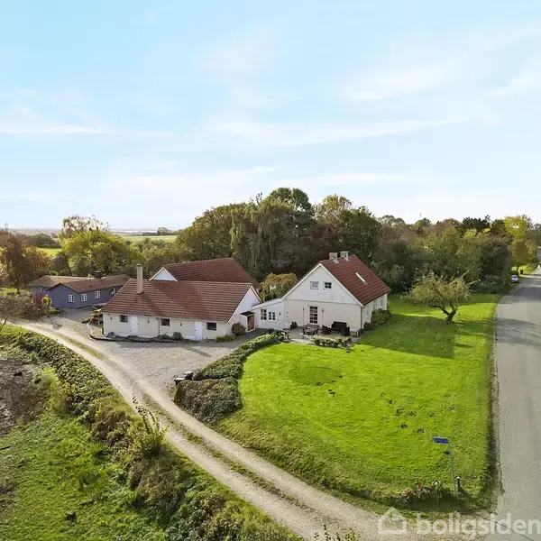 Et landhus med røde tage står ved en grusvej. Handlingen viser huset omkranset af græsplæne og træer. Konteksten er et landligt område med en asfaltvej til højre.