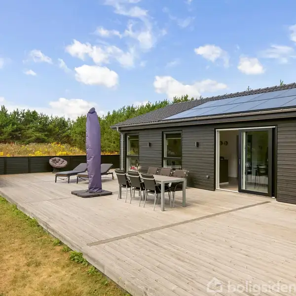 Scandinavisk sommerhus med terrasse, hvor et havemøbelsæt står under en stor parasol. Huset har solpaneler på taget. Omkring huset er der græs og træer.
