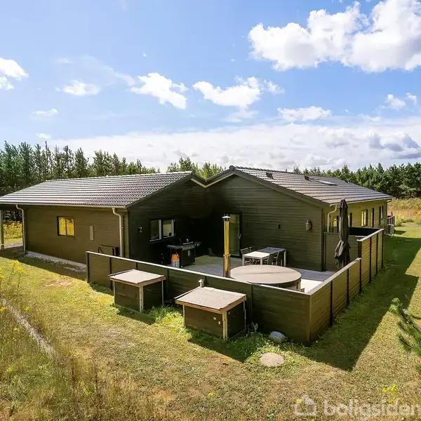 Træhus står stille omgivet af græs og træer. Terrasse med borde og stole ses foran huset. Sollys skinner gennem en let skyet himmel.