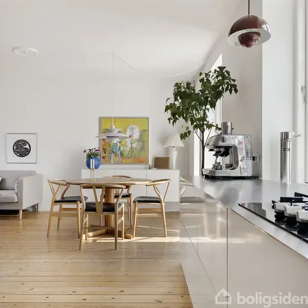 Rundt spisebord med fire stole står i moderne, lyst køkken. Espressomaskine ses ved vindue. Væggen prydes af kunstværker, og en stor potteplante giver rummet liv.