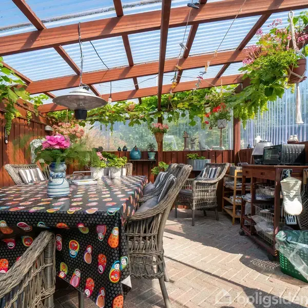 En overdækket terrasse med et bord dækket af en farverig dug og omgivet af kurvestole. Over hænger planter fra træbjælkerne. Hylder langs væggen med forskellige genstande.